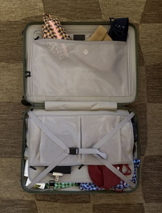 Open suitcase on a patterned carpet, packed with clothes, accessories, and toiletry bags. Mesh dividers and straps secure the items inside.