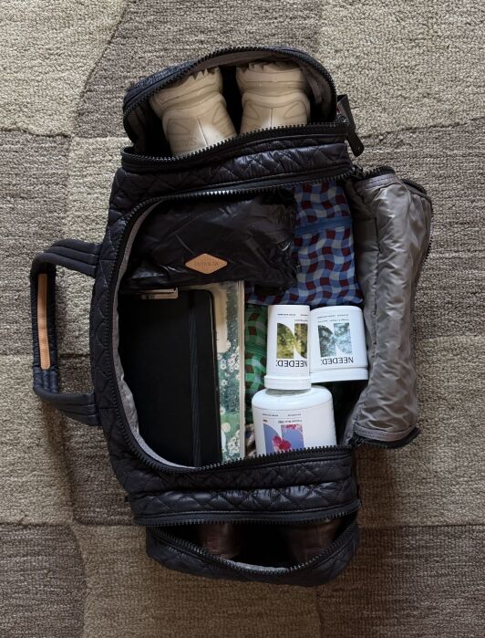 An open black duffel bag showing white sneakers, clothes, a tablet, two supplement bottles, and a folded jacket inside, placed on a beige and brown patterned carpet.