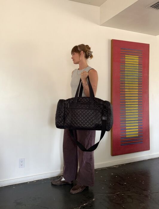 Person standing indoors holding a large black quilted duffel bag, wearing a sleeveless top and wide-leg pants, with a colorful abstract painting on the wall in the background.