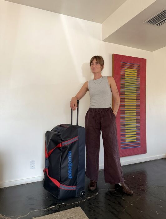 A person stands indoors beside a large black Patagonia rolling suitcase, wearing a gray sleeveless top and brown pants, with a colorful artwork on the wall behind them.