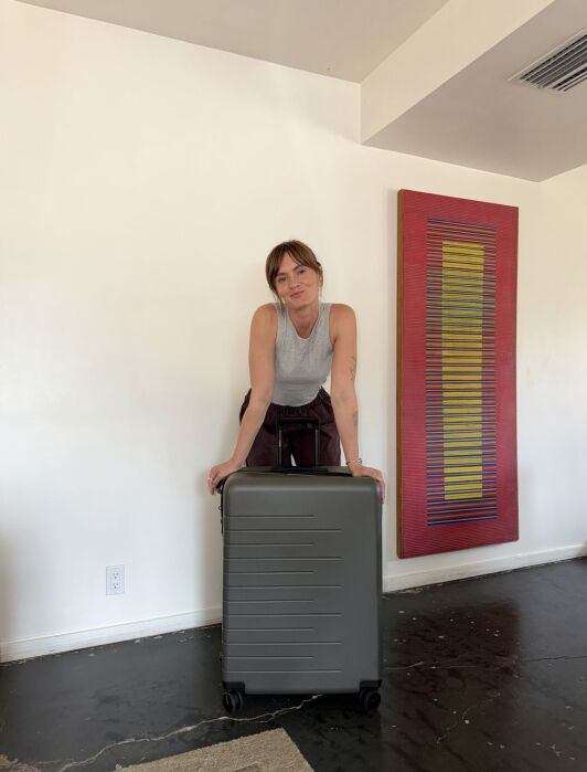 A woman stands indoors, leaning on a large gray suitcase. The room has white walls, dark floors, and a colorful abstract painting on the wall.