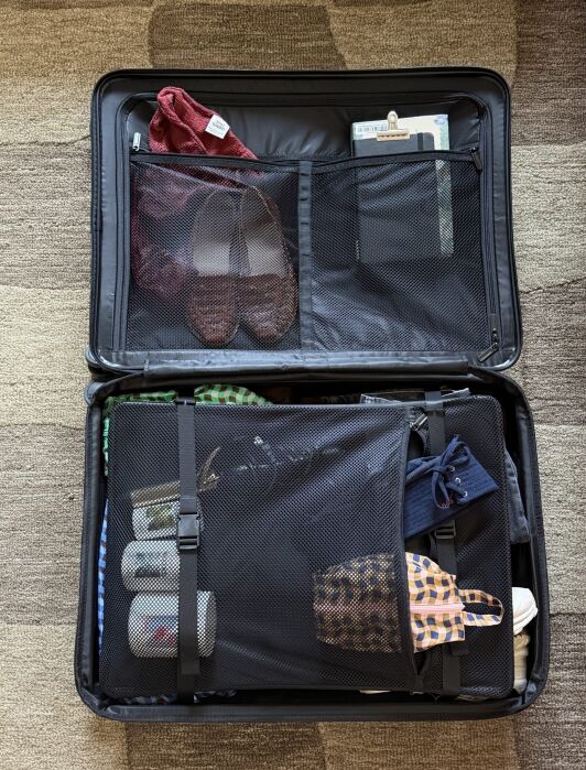 Open suitcase on a patterned carpet, neatly packed with clothes, shoes, toiletries, a face mask, and a book in mesh compartments and straps.