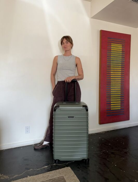 A person stands indoors next to a large green suitcase, resting one hand on the handle. A colorful abstract painting hangs on the wall.