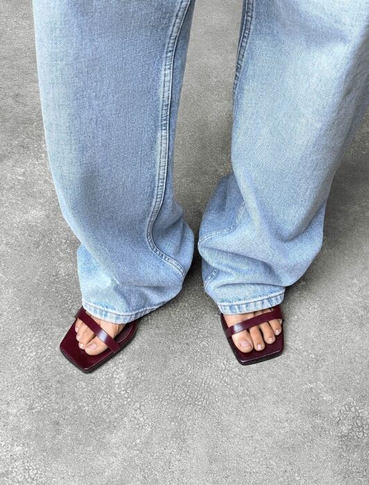 Person wearing light blue jeans and burgundy square-toed sandals, standing on a textured gray surface.