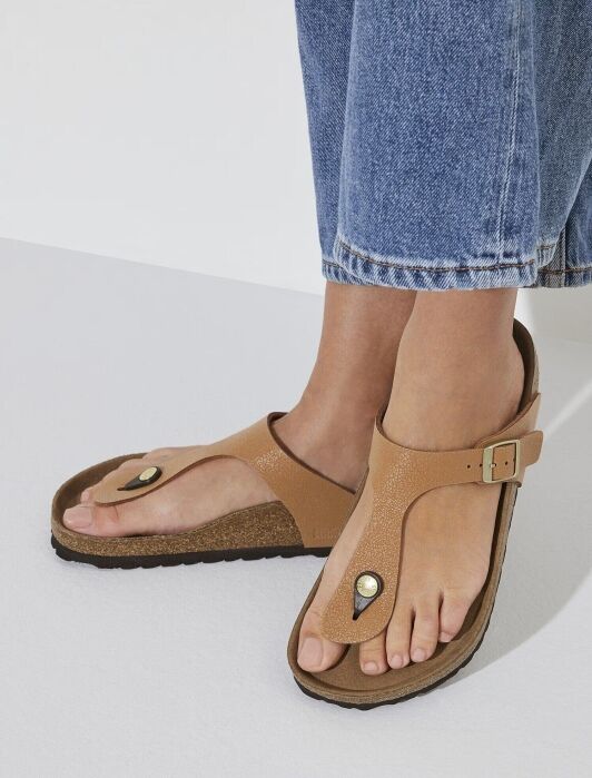 A person wearing tan thong sandals with a buckle strap, standing on a white surface and dressed in cropped blue jeans.