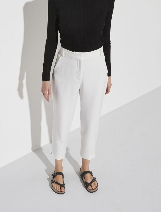 Person wearing a long-sleeve black top, white high-waisted cropped pants, and black sandals standing against a plain white background.