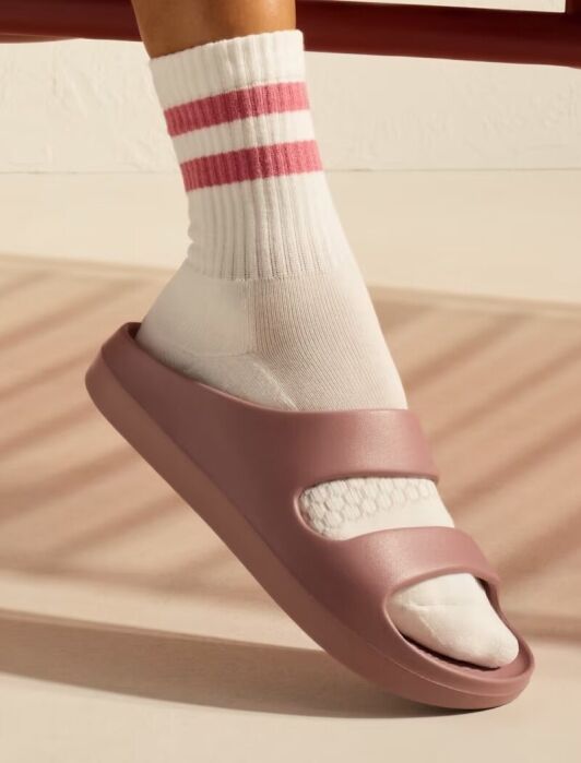 A person wearing a white sock with pink stripes and a mauve slide sandal stands on a light-colored floor with a shadow falling across it.