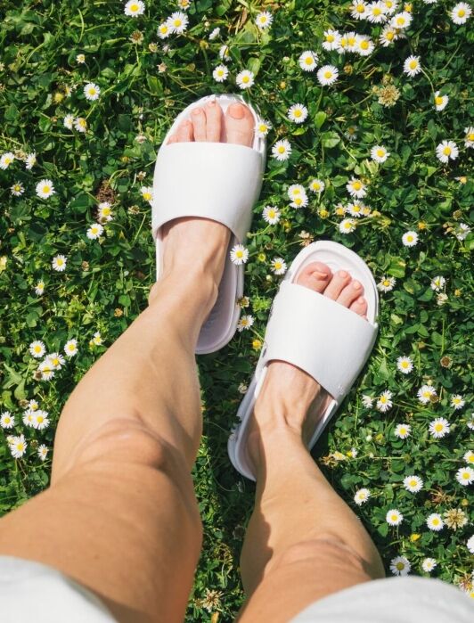 Person wearing white sandals standing on grass with small white daisies.