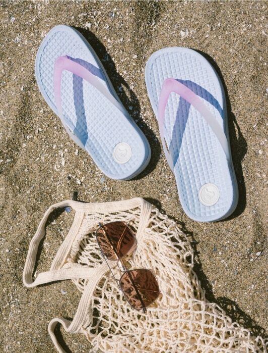 A pair of blue and purple flip-flops, sunglasses, and a netted bag lying on sandy beach.
