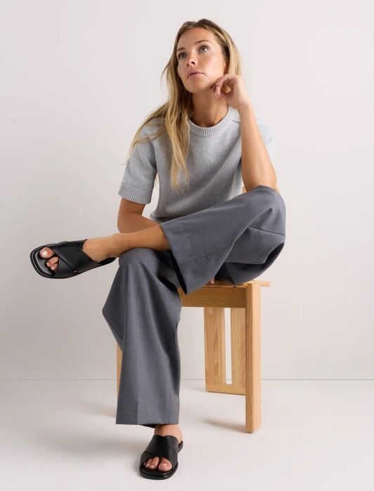 A woman sits on a wooden stool wearing a light grey short-sleeve sweater, grey wide-leg pants, and black slide sandals, with one leg crossed over the other.