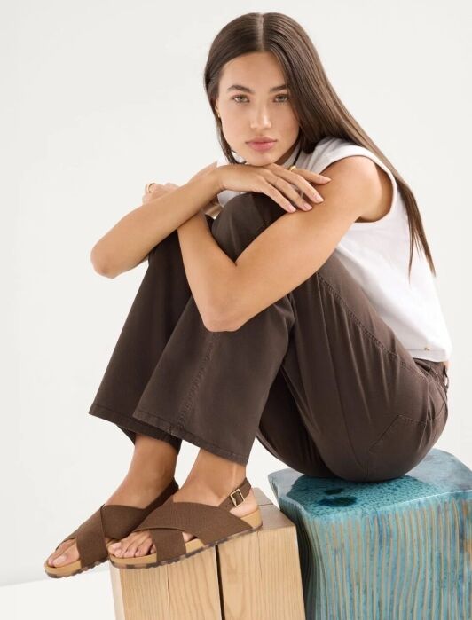 Woman with long dark hair sits on a blue wooden block, wearing a white sleeveless top, brown pants, and brown sandals, with arms resting on her knees.