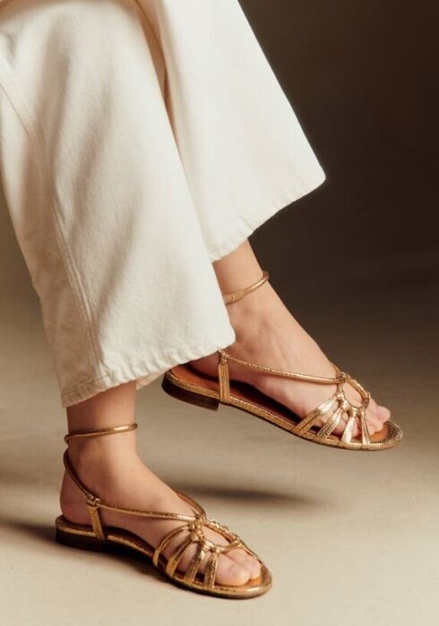 A person wearing wide-leg white pants and gold strappy flat sandals stands against a neutral background.