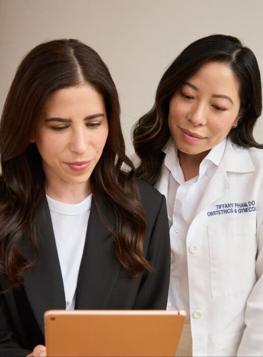 Two women stand together looking at a tablet; one wears a black blazer and the other a white medical coat that reads "Obstetrics & Gynecology.