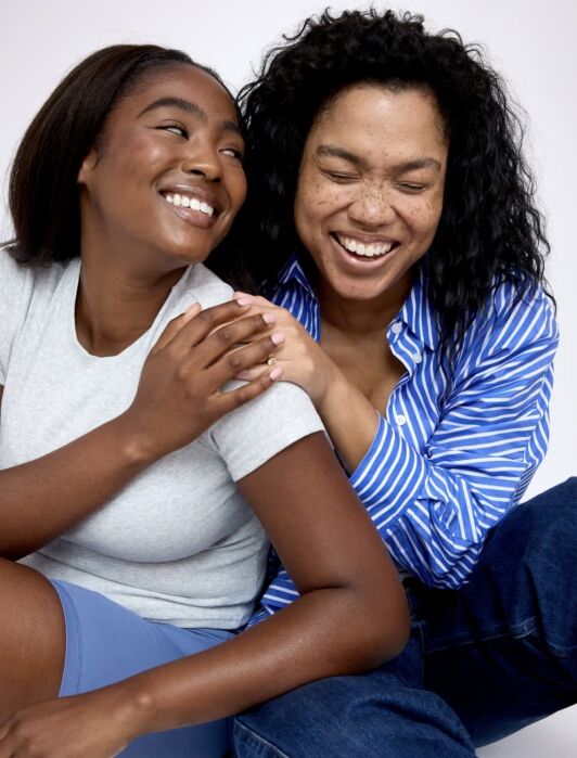 Two people sitting close together, smiling and laughing. One is wearing a light grey shirt, the other a blue striped shirt. The person in blue rests their hand on the other's shoulder.