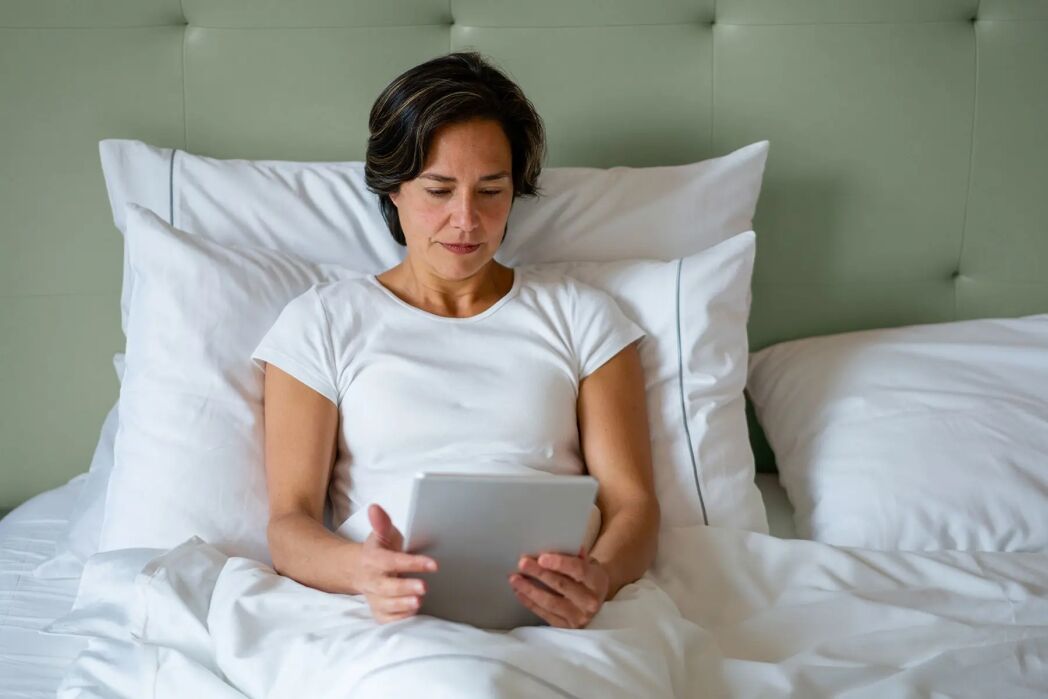 A woman in a white shirt sits up in bed with white bedding, looking at a tablet device.