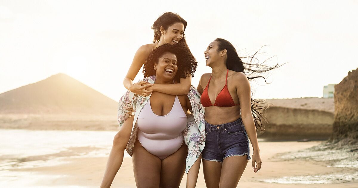 Three women in swimsuits and shorts laugh and walk together on a beach at sunset; one woman gives another a piggyback ride.