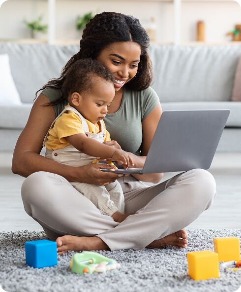 A woman sits cross-legged on the floor with a baby on her lap, both looking at a laptop. Colorful toys are scattered on the rug in front of them.