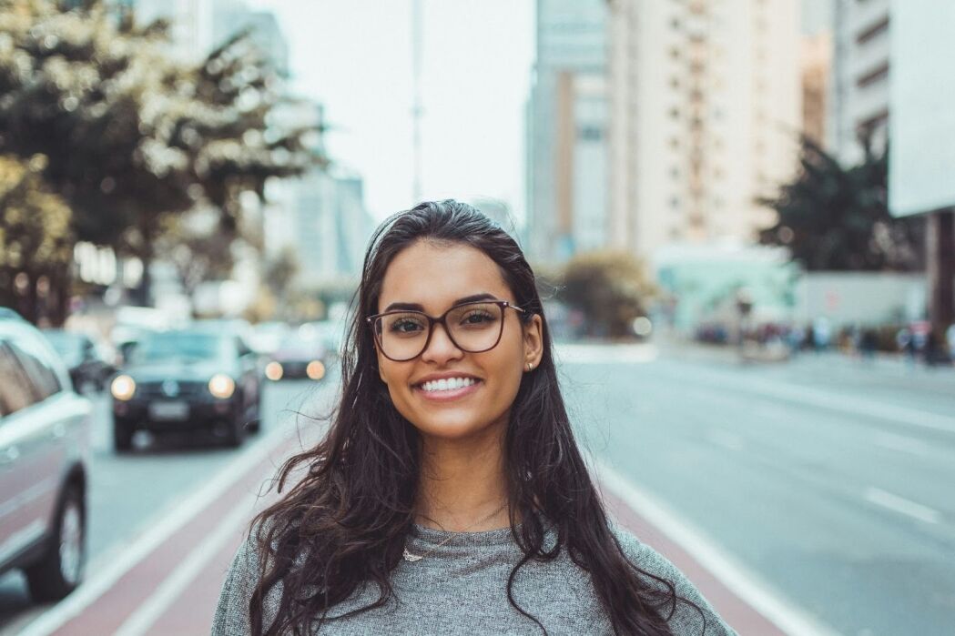 A woman with long dark hair and glasses smiles at the camera while standing on a city street with buildings and cars in the background.