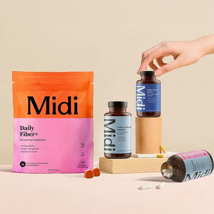 A hand reaches for a bottle among various Midi supplement products, including a colorful fiber pouch, pill bottles, capsules, and gummies arranged on a beige surface.