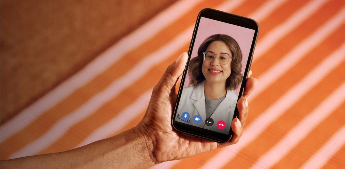 A person holds a smartphone displaying a video call with a woman wearing glasses and a white coat, suggesting a virtual consultation.