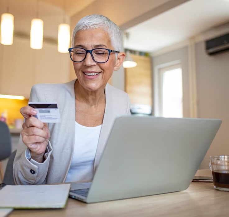 Older woman with short gray hair and glasses sits at a table using a laptop, holding a credit card, with a notebook and a glass of coffee nearby.