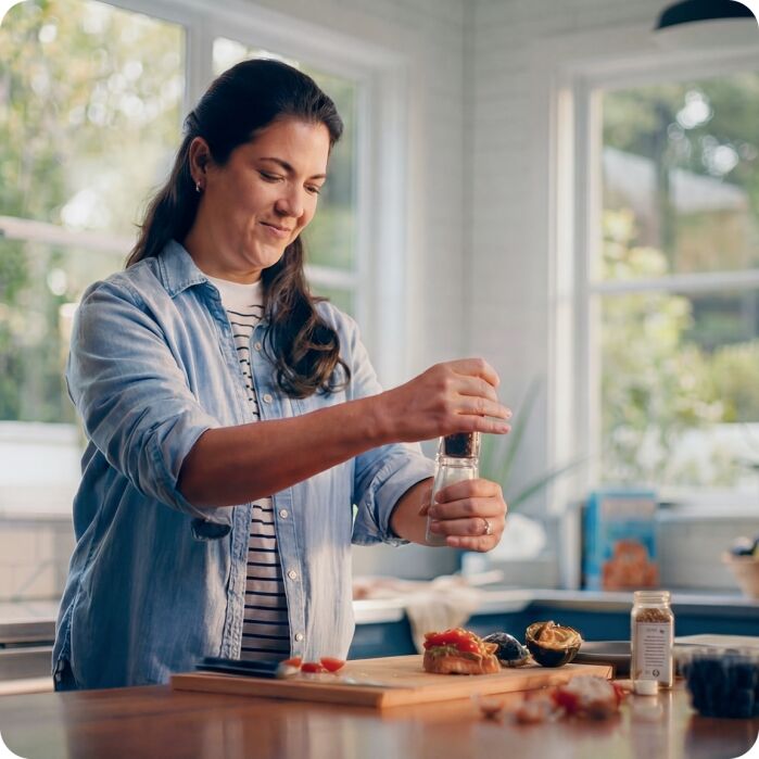 A woman in a kitchen grinds pepper onto food on a cutting board, with various ingredients and a jar of spices visible nearby.