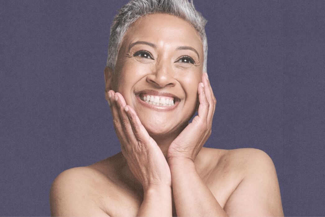 Smiling older person with short gray hair holds their face with both hands against a plain purple background.
