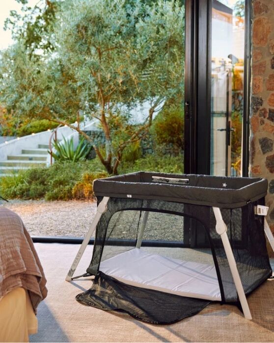 A black and white portable baby crib is set up indoors near a large glass door, with a garden visible outside.