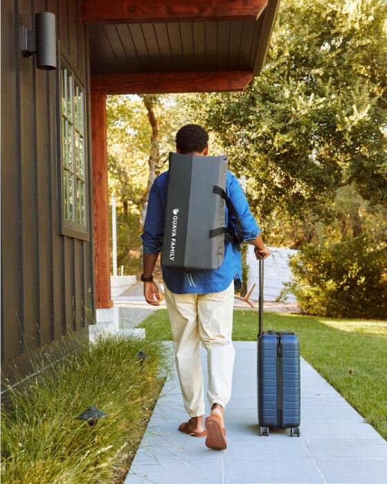 A person walks on a path carrying a yoga mat on their back and pulling a blue suitcase, next to a dark building with trees in the background.