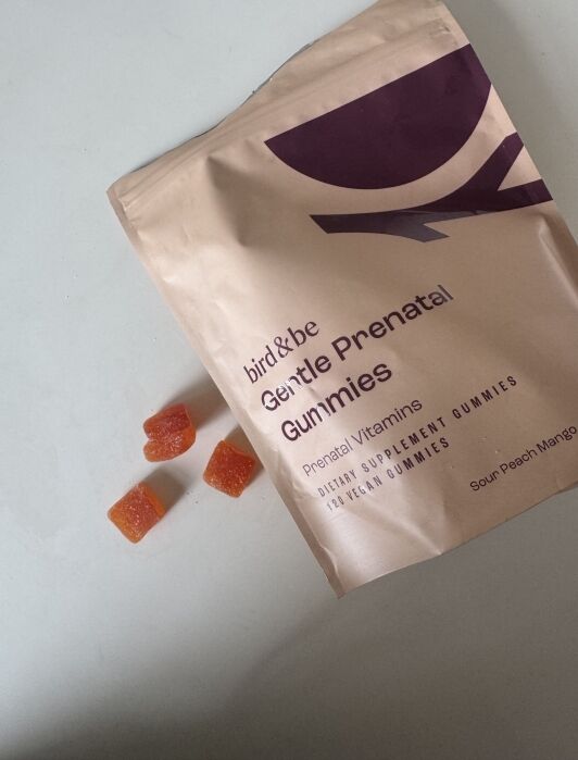A package labeled "bird&be Gentle Prenatal Gummies" with four orange prenatal vitamin gummy squares beside it on a white surface.