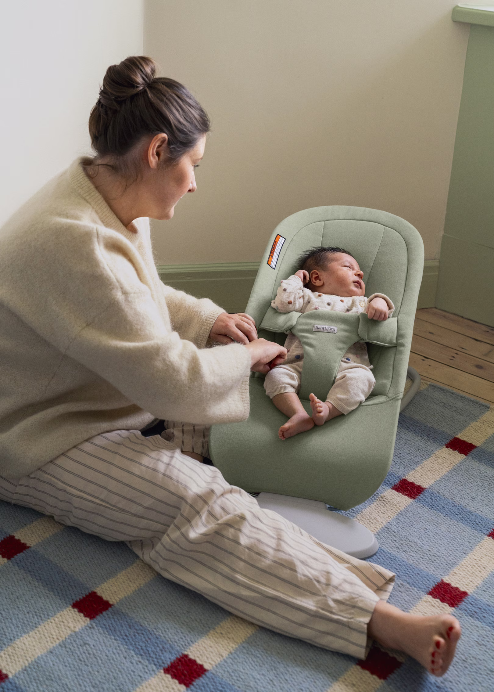 Woman sits on the floor and adjusts a green baby bouncer with an infant lying inside, on a blue and red plaid rug in a softly lit room.