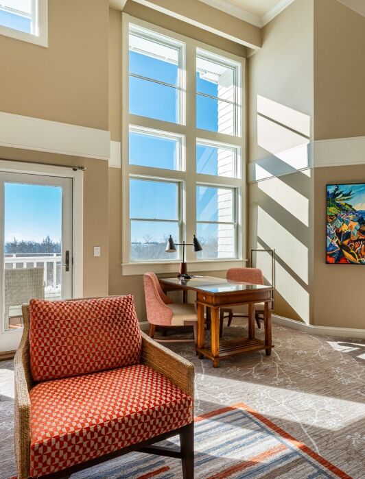 A bright room with large windows, beige walls, a red armchair, wooden desk with two pink chairs, and a colorful painting on the wall. Sunlight streams through the windows.