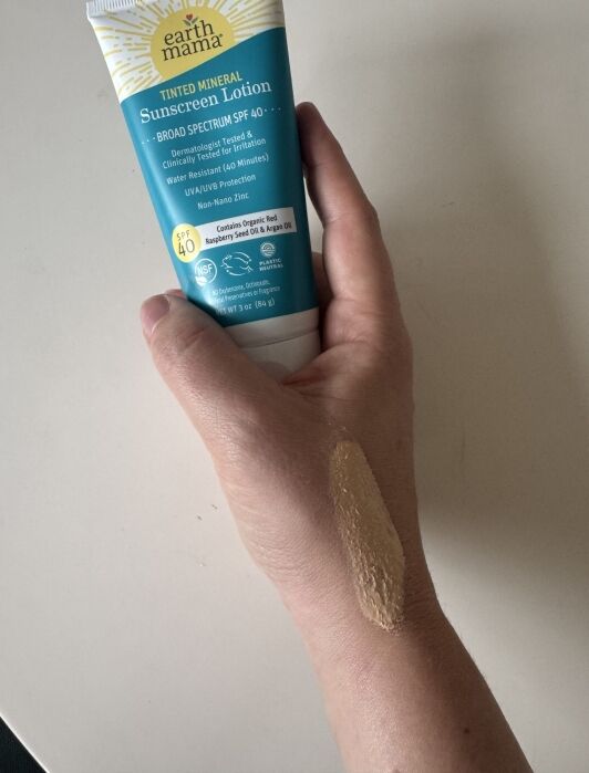 A hand holds a tube of Earth Mama Tinted Mineral Sunscreen Lotion, with a streak of the lotion applied to the back of the hand.