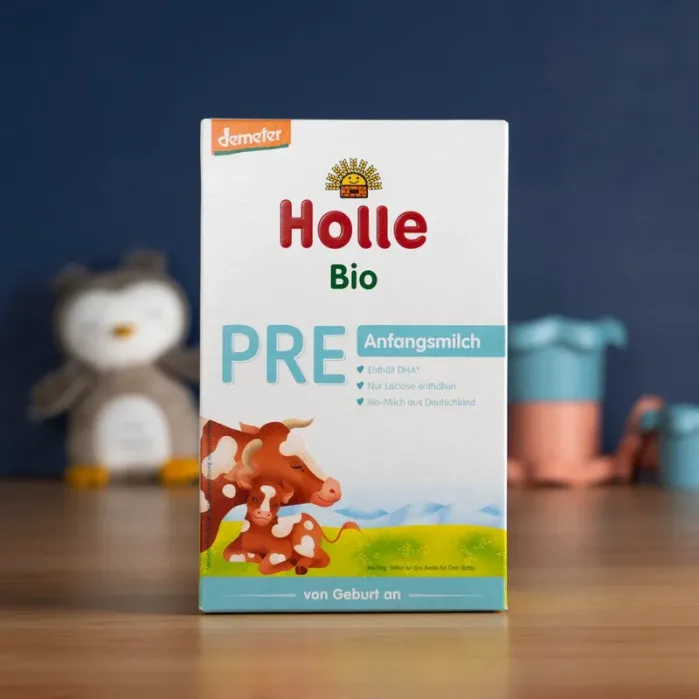 Box of Holle Bio PRE infant formula sits on a wooden surface, with a blurred background showing a toy owl and other objects.