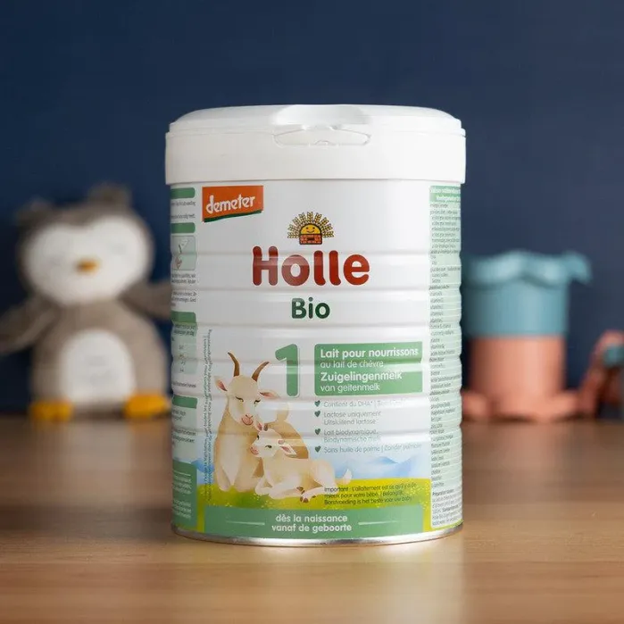 A can of Holle Bio Stage 1 organic infant formula sits on a table with plush toys and a cup in the blurred background.