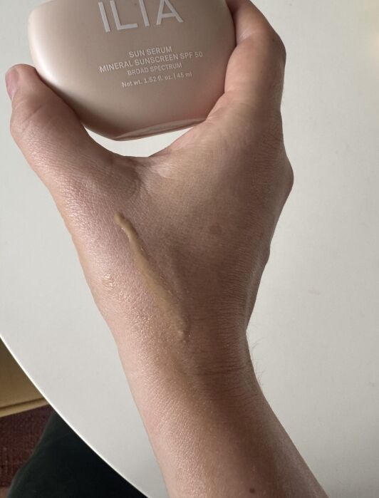 A hand with a streak of beige sunscreen on the back holds a bottle labeled "ILIA Sun Serum Mineral Sunscreen SPF 50.