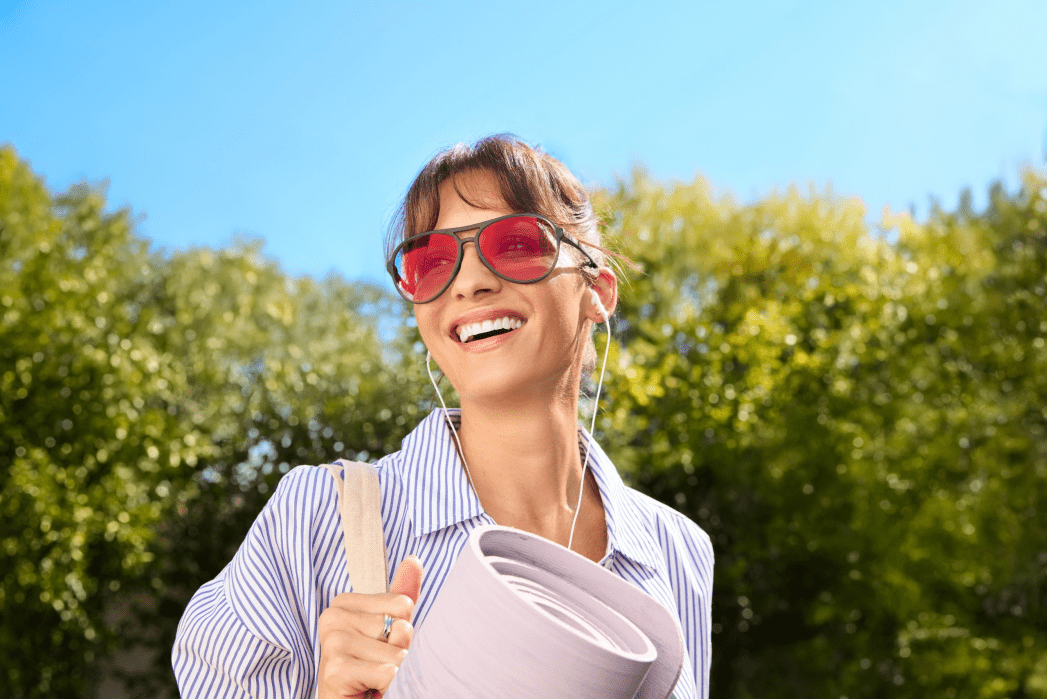 Woman wearing pink sunglasses and earphones, carrying a rolled-up yoga mat, smiling outdoors with trees and blue sky in the background.