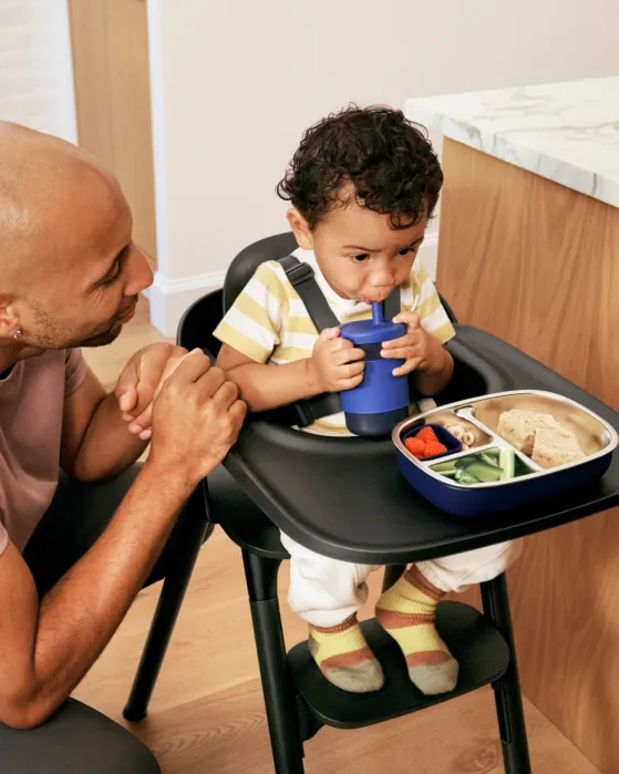 A toddler sits in a high chair drinking from a cup, with a divided plate of food in front. An adult kneels beside the chair, watching the child.