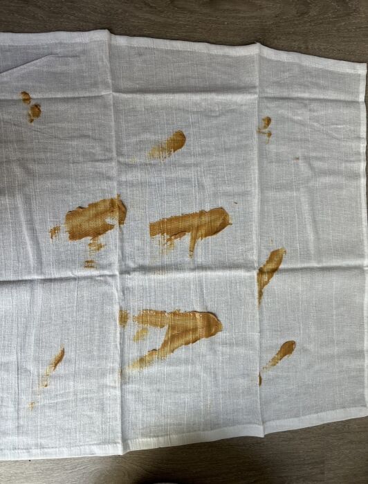 A white cloth napkin is spread out on a wooden surface, showing several brown-orange stains and smudges in various areas.