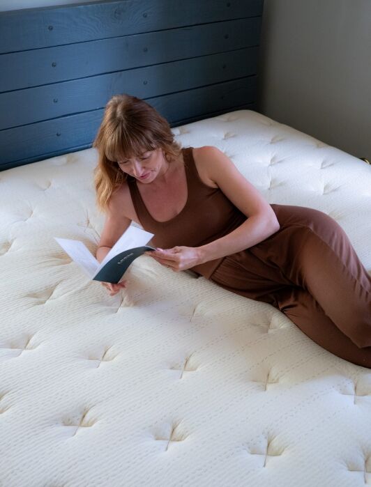 Woman in a brown outfit lies on a white mattress, reading a booklet, with a wooden headboard in the background.