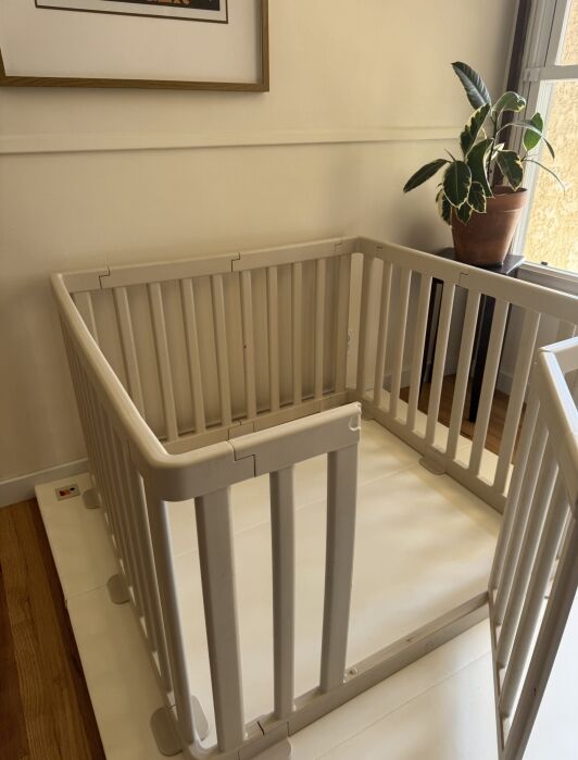 A beige baby playpen is set up in the corner of a room near a window with a potted plant on the windowsill and framed art on the wall.
