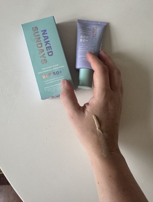 A hand with sunscreen swatched on the back is shown next to a Naked Sundays SPF 50+ sunscreen tube and its box on a white surface.