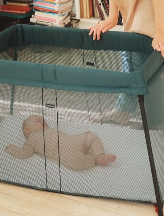 A baby lies in a mesh-sided playpen on a white mattress while an adult stands nearby. Books are stacked on shelves in the background.
