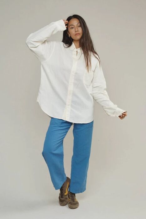A person stands against a plain background wearing a loose white button-up shirt, blue pants, and brown shoes, with one hand touching their head.