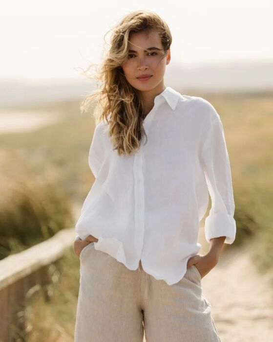 A person stands outdoors on a sandy path wearing a white button-up shirt and light-colored pants, with one hand in their pocket.