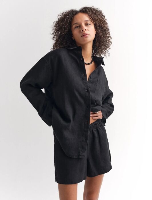 A person with curly hair wears a black button-up shirt and matching black shorts, standing against a plain white background.
