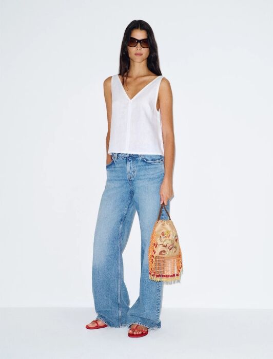 Woman standing against a white background, wearing a white sleeveless top, wide-leg blue jeans, red sandals, sunglasses, and holding an embroidered bag with orange fringe.