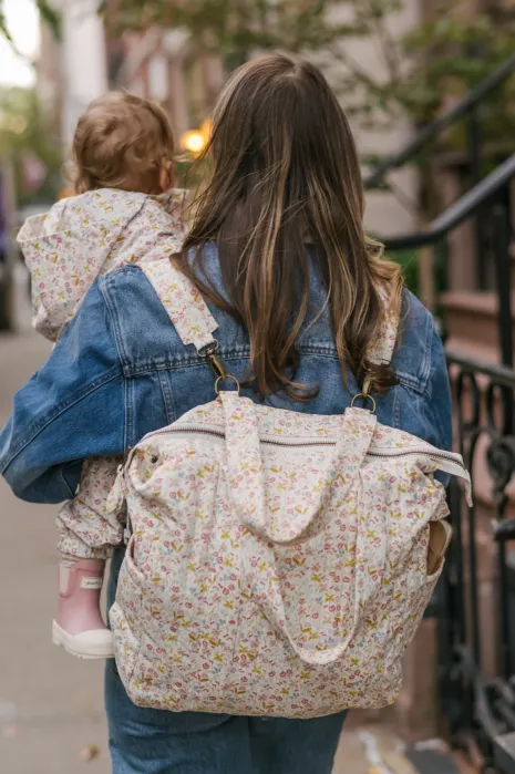 Woman with long hair carries a toddler and a floral diaper bag while walking outdoors on a city sidewalk. Both wear floral-patterned clothing.