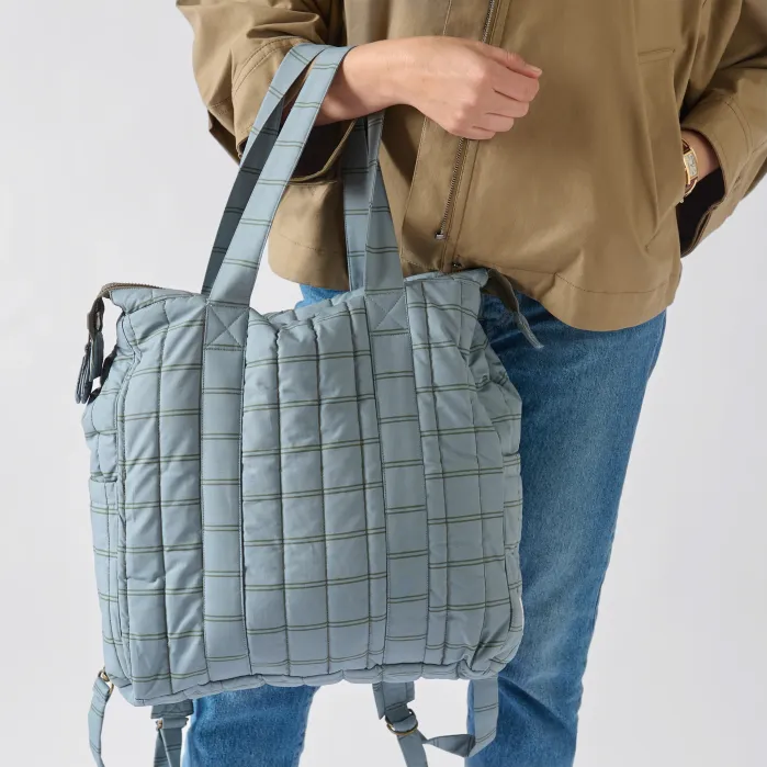 Person wearing a tan jacket and blue jeans holds a large, quilted light blue bag with double handles and backpack straps.