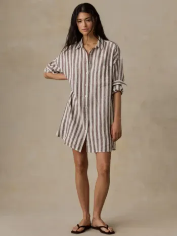 A person stands against a plain background wearing a brown and white vertically striped, long-sleeve button-down shirt dress and black sandals.
